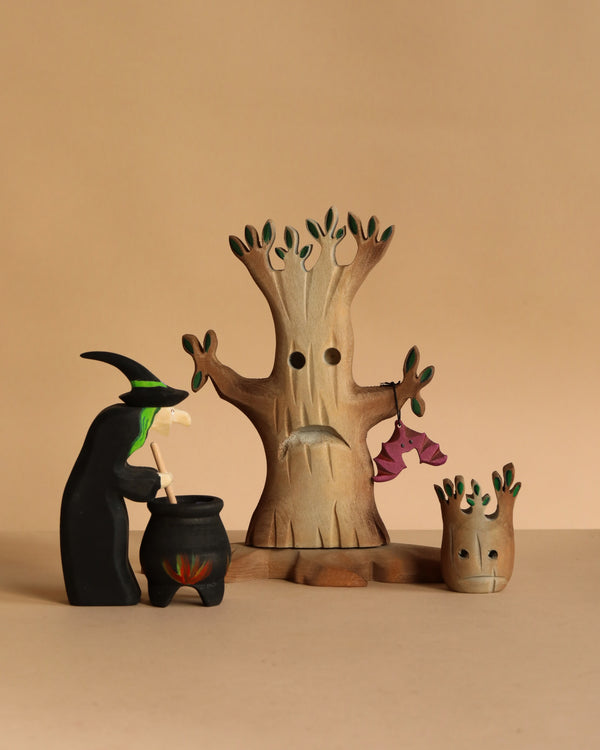 The Bumbu Handmade Witch with Cauldron and Haunted Tree Set features a wooden witch stirring her cauldron beside two tree figures, creating a charming Montessori Waldorf display against a simple beige background.