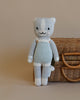 A handmade, hand-knit Cuddle + Kind Cat Stuffed Animal in striped overalls standing in front of a wicker basket on a neutral background. The bear exhibits a smiling face and is crafted in light gray and white.