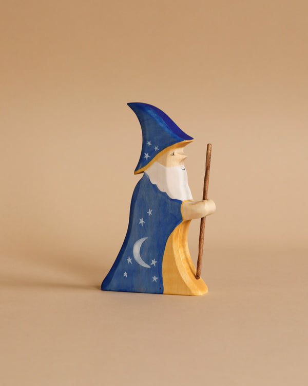 A Handmade Wooden Wizard, meticulously crafted from sustainable hardwood, is featured against a beige background. The wizard, painted in non-toxic blue, yellow, white, and brown colors, boasts a long white beard and wears a dark blue pointed hat and robe adorned with star and moon designs while holding a staff.