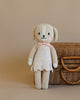 Cuddle + Kind Mia the Dog is a hand-knit plush made from premium cotton yarn, featuring a light pink dress and bow. She stands in front of a brown wicker basket on a beige background.