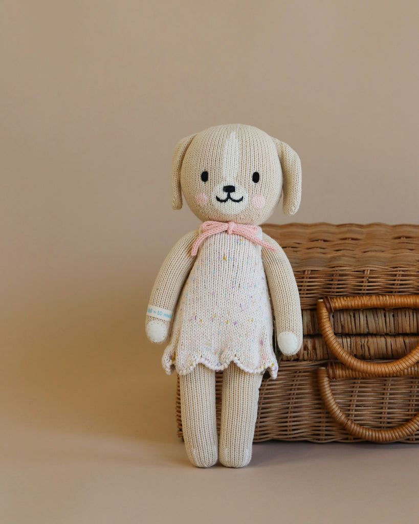 A Cuddle + Kind Dog Stuffed Animal with a smiling face, wearing a pastel pink scarf, stands upright next to a woven basket on a light brown background.