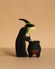 The Bumbu Handmade Witch with Cauldron and Haunted Tree Set features a small wooden witch with green hair and a black hat stirring a flame-adorned cauldron, displayed against a plain beige background.