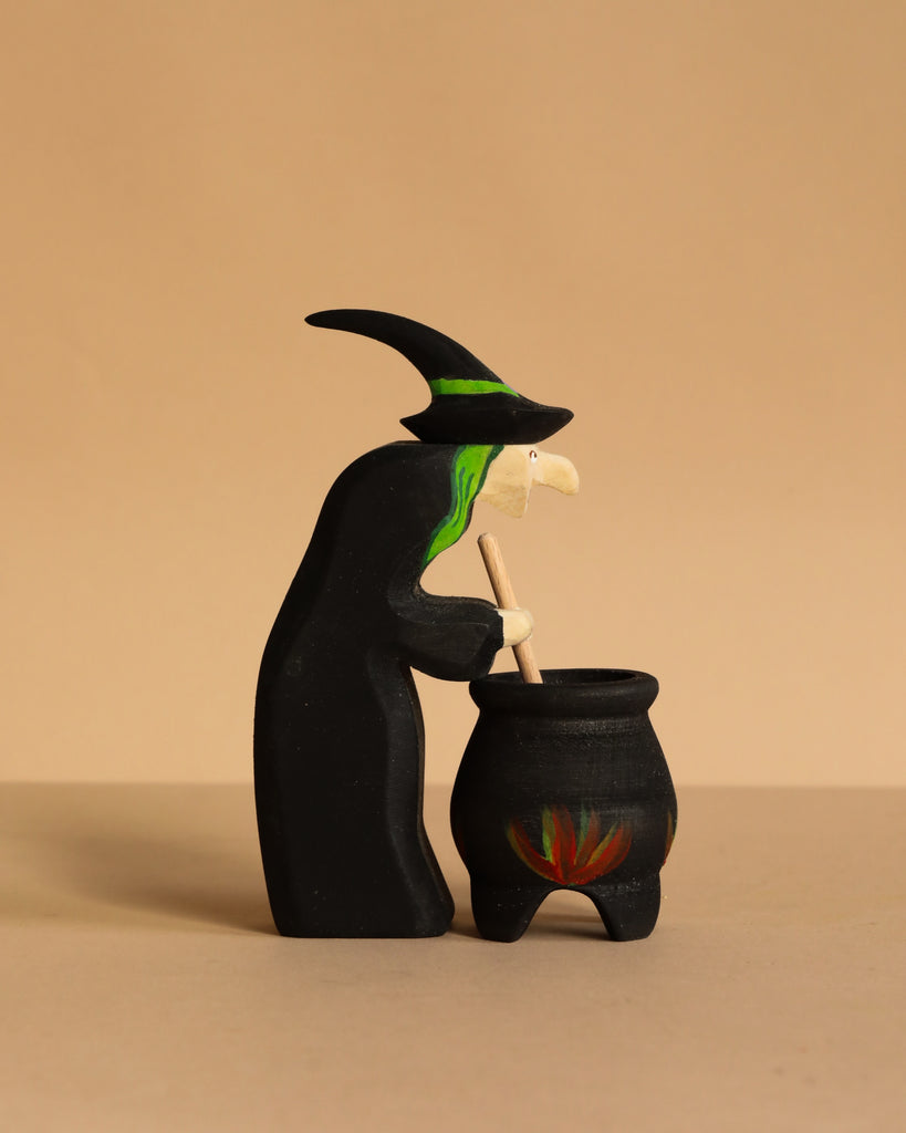 The Bumbu Handmade Witch with Cauldron and Haunted Tree Set features a small wooden witch with green hair and a black hat stirring a flame-adorned cauldron, displayed against a plain beige background.