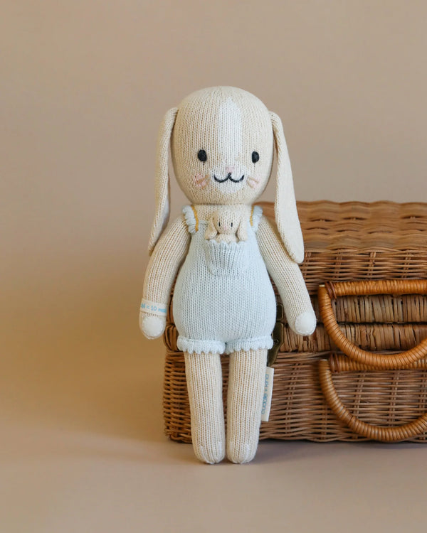 Cuddle + Kind Briar the Bunny - Light Blue is a hand-knit cotton doll dressed in light blue overalls, holding a tiny toy bunny and standing in front of a closed wicker basket on a beige background.