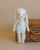 Cuddle + Kind Briar the Bunny - Light Blue is a hand-knit cotton doll dressed in light blue overalls, holding a tiny toy bunny and standing in front of a closed wicker basket on a beige background.