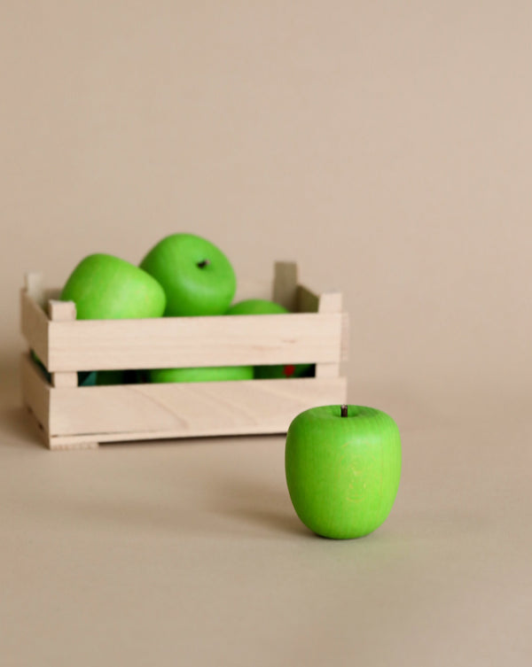 A wooden crate filled with Erzi Apple Green Pretend Food miniature green apples on a beige background, with one apple placed in front of the crate.