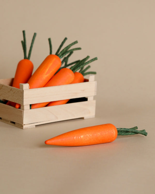 A wooden box filled with Erzi Carrot Pretend Food, specifically fresh orange carrots with green tops, handcrafted in Germany, placed on a neutral beige background. One carrot lies outside the box in the foreground.