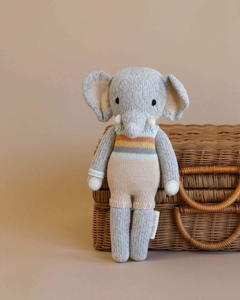 A Cuddle + Kind Elephant Stuffed Animal, featuring a gray body with white accents and a colorful striped sweater filled with hypoallergenic polyfill, sits on a wicker basket against a soft beige background.