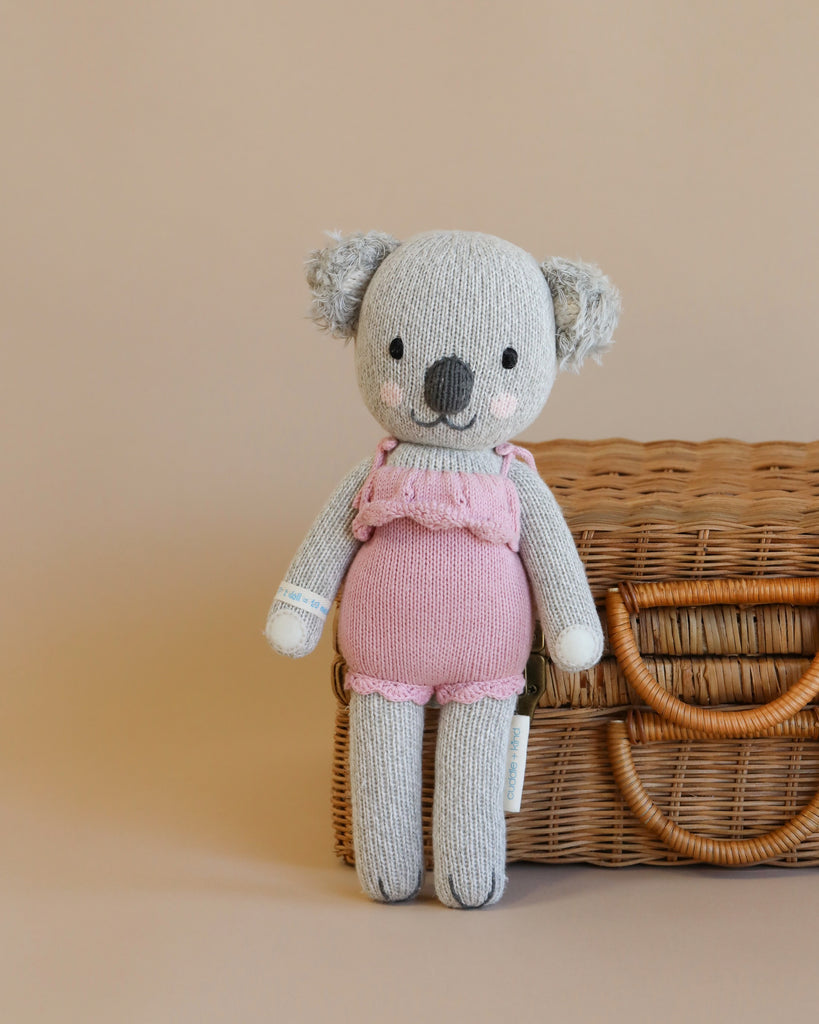 A hand-knit Cuddle + Kind Koala Stuffed Animal in a pink outfit sitting on a wicker basket against a light beige background. The toy has a smiling face and wears a cozy hat.