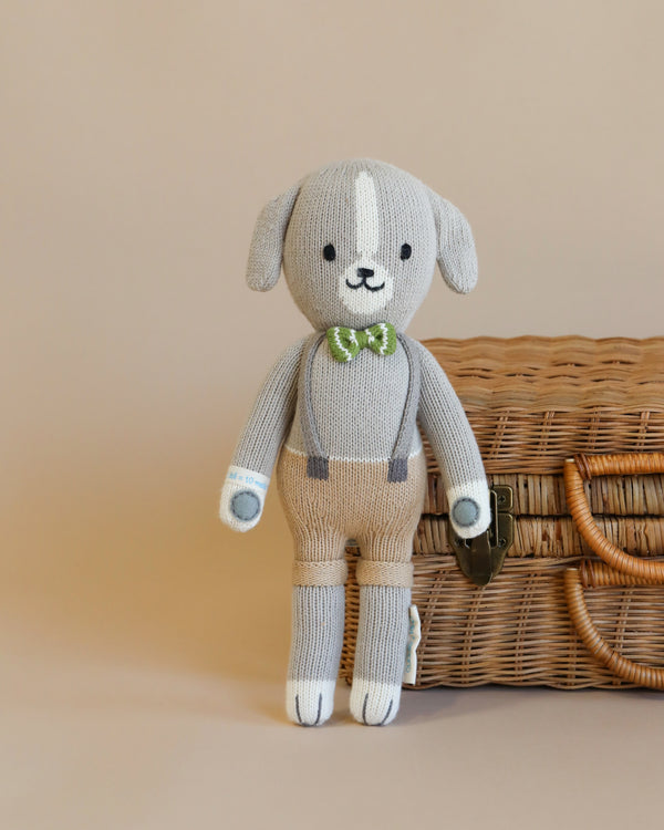Cuddle + Kind Noah the Dog is a hand-knit gray and cream stuffed animal wearing a green bow tie and suspenders, posed before a woven wicker basket with a beige background.