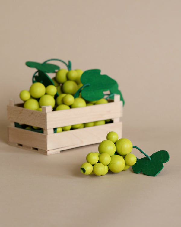 A wooden crate filled with Erzi Green Grapes Pretend Food, made from beechwood beads, alongside two grape bunch shapes, on a beige background. One grape bunch is placed next to the crate.