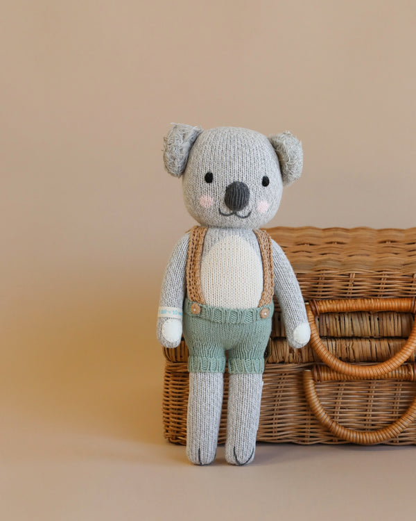 Cuddle + Kind Quinn the Koala, a hand-knit doll with pink cheeks and green shorts with brown suspenders, stands before a closed wicker basket on a beige background.