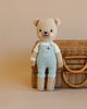 A Cuddle + Kind the Honey Bear in light blue overalls standing in front of a wicker basket on a neutral background.
