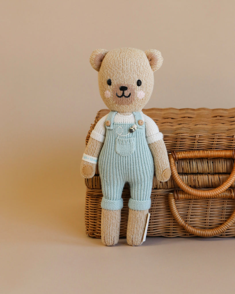 A Cuddle + Kind the Honey Bear in light blue overalls standing in front of a wicker basket on a neutral background.