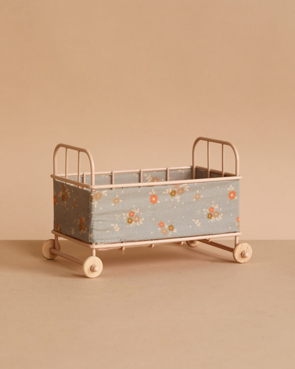 The Maileg Cot Bed, Micro - Powder features a light pink metal frame with wheels and blue floral fabric sides, and is shown on a beige surface against a matching beige background.