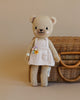 A Cuddle + Kind  the Honey Bear in a white dress stands beside a wicker basket on a beige background. The bear has a small bouquet of flowers in its pocket and is stuffed with hypoallergenic.