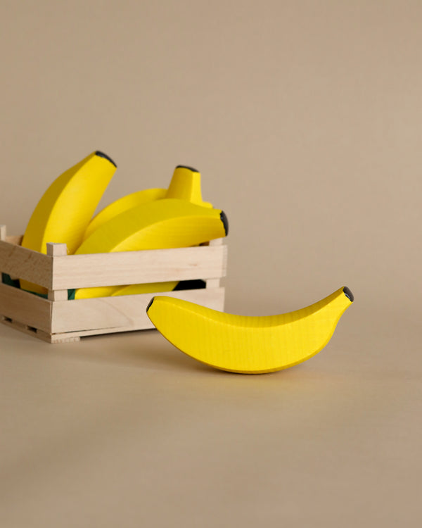 A wooden crate filled with yellow wooden bananas from Erzi Banana Pretend Food, with one banana lying in front of the crate on a beige surface.