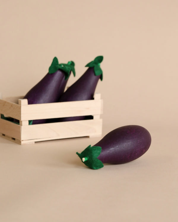 A small wooden crate containing several Erzi Eggplant Pretend Foods, with one Erzi Eggplant Pretend Food lying in front of it, all against a plain, light beige background.