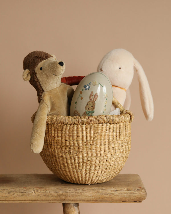 The Easter Basket Set, with a wicker basket on wood, cradles a Senger Naturwelt stuffed hedgehog, a stuffed rabbit, and a rabbit-decor egg. The soft beige background enhances this handmade creation's charm.