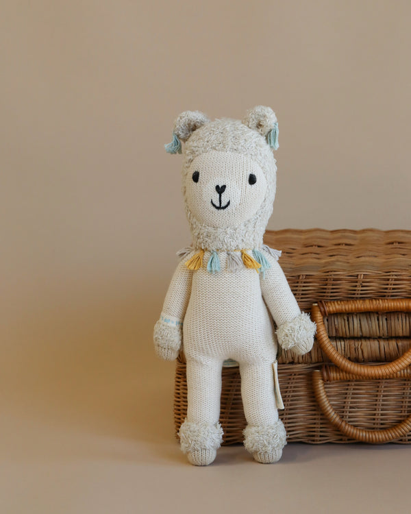 Cuddle + Kind Lucas the Llama, a hand-knit, hypoallergenic stuffed doll with small ears, friendly face, and pastel accents, sits upright by a wicker basket against a neutral background—adorable and perfect for all ages.