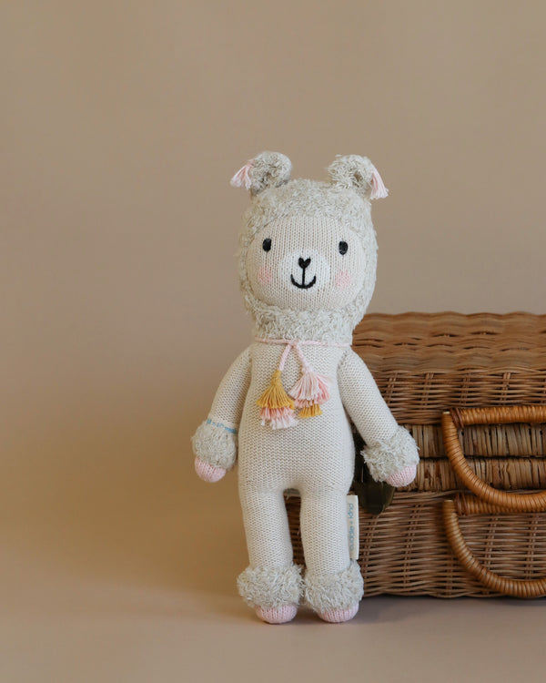 The Cuddle + Kind Lola the Llama, a hand-knit doll with pink cheeks and a sweet smile, stands before a closed wicker basket on a simple beige background.