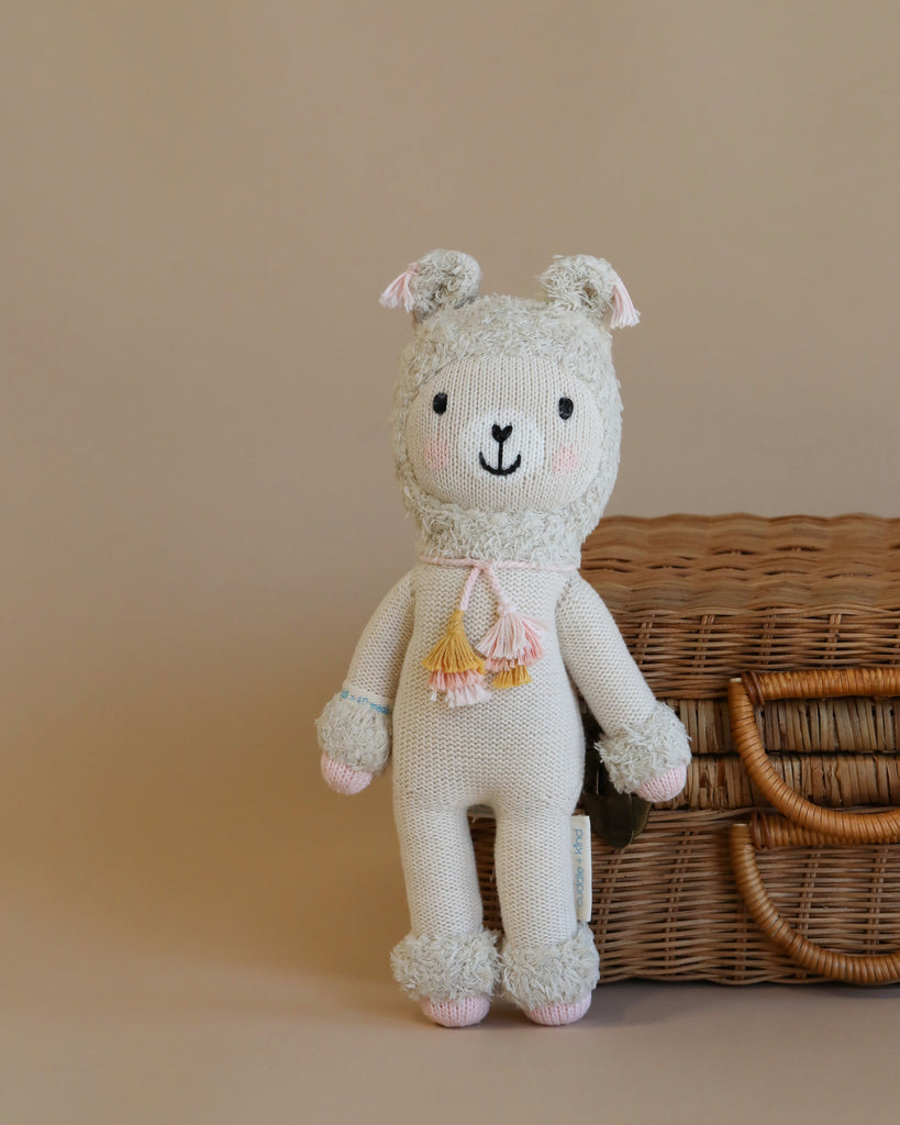 A Cuddle + Kind Llama Stuffed Animal with pink accents and a tassel belt, filled with hypoallergenic polyfill, standing in front of a wicker basket, against a beige background.