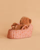 The Maileg Teddy Bear in Carry Cot features a soft brown plush bear resting in a pink floral-patterned cot with ribbon handles, displayed on a plain peach background.