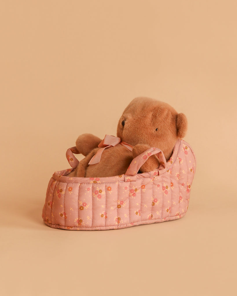 The Maileg Teddy Bear in Carry Cot features a soft brown plush bear resting in a pink floral-patterned cot with ribbon handles, displayed on a plain peach background.