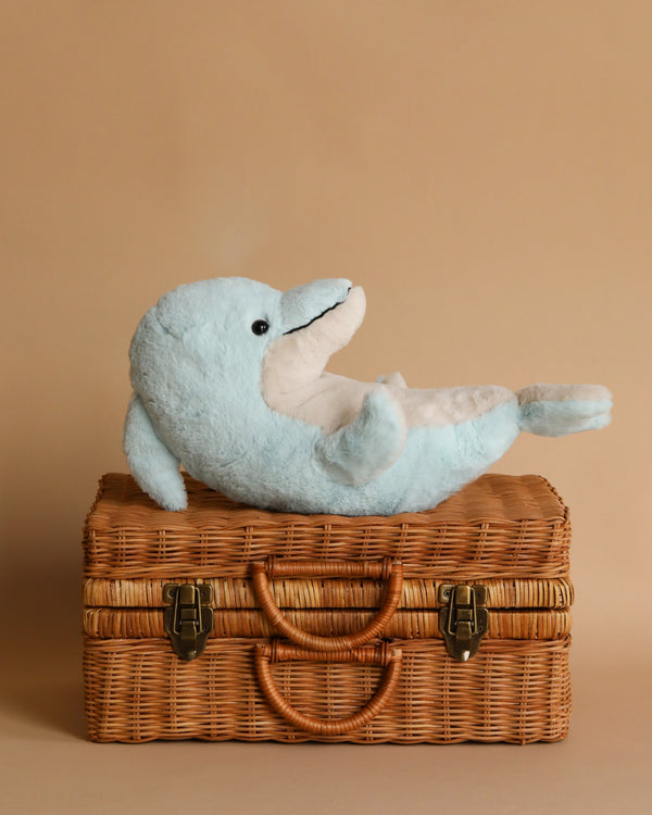 A plush blue Steiff, Dolphin Stuffed Plush Animal toy reclining on a brown wicker suitcase against a warm beige background, creating a playful yet serene display.