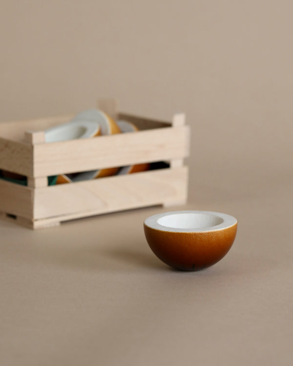 A small, Erzi Coconut Half Fruit resembling a realistic half coconut with a white interior, placed in the foreground on a beige surface, with a wooden crate containing similar bowls blurred in the background.