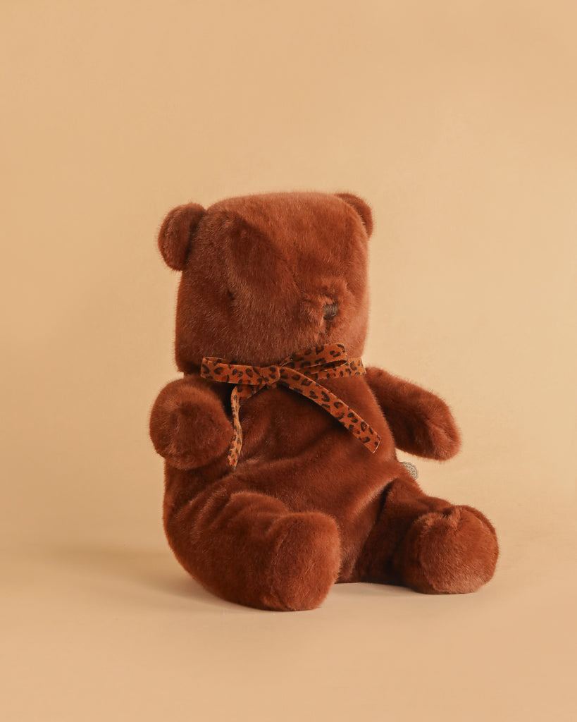 The Maileg Teddy Bear Plush, Medium - Dark Chocolate, sits upright on a beige surface with soft brown fur, minimal facial features, and a leopard print ribbon around its neck for a charmingly cozy look.