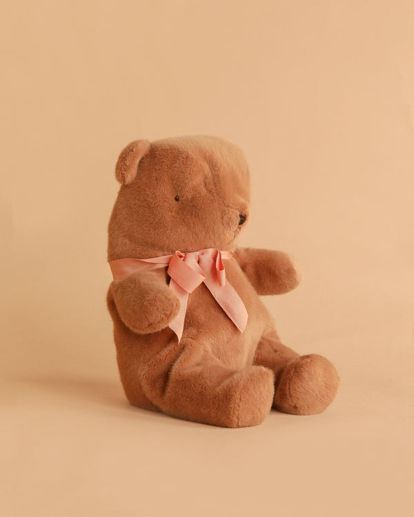 The Maileg Teddy Bear Plush, Medium - Soft Brown, sits upright on a beige background with a pink bow around its neck, exuding a charming vintage vibe.
