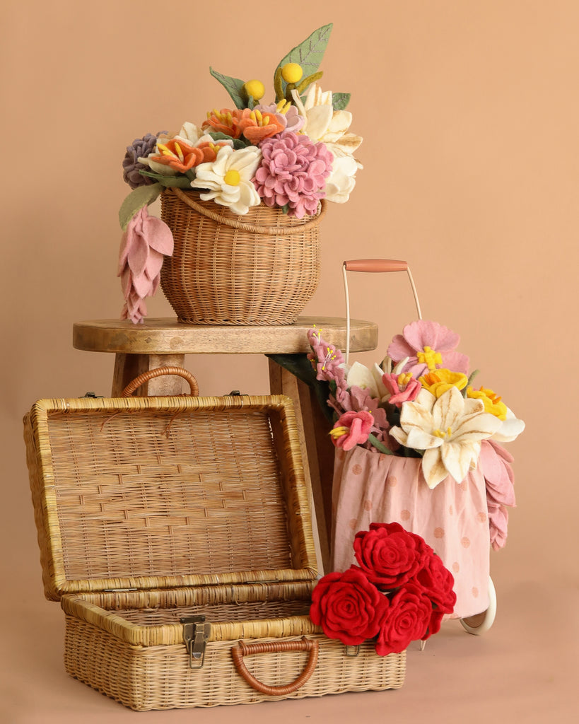 The Basket of Felt Flowers, featuring vibrant felt blooms in three wicker baskets—one open to reveal its empty interior—is displayed on a wooden stool and floor with red felt roses arranged in front against a beige background.