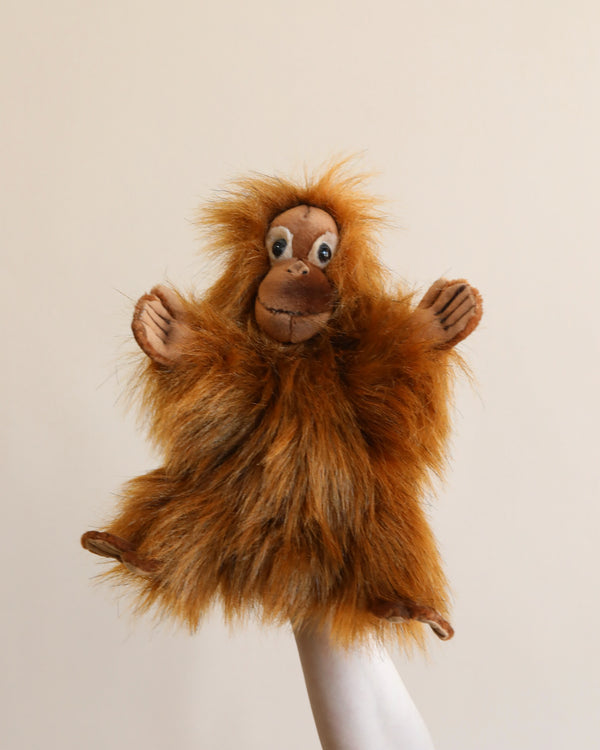 A whimsical Orangutan Puppet with shaggy brown fur and expressive eyes perched playfully on a human hand against a light beige background. This HANSA animal crafted to be lifelike