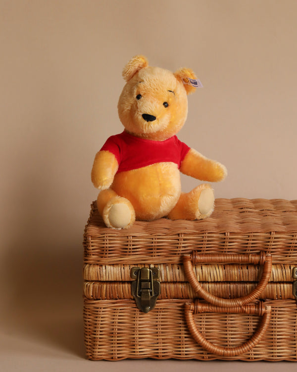 A Steiff Disney's Winnie the Pooh Open Edition Collectible, 10 Inches toy sits atop a wicker suitcase against a neutral background, dressed in a red shirt and looking cheerful.