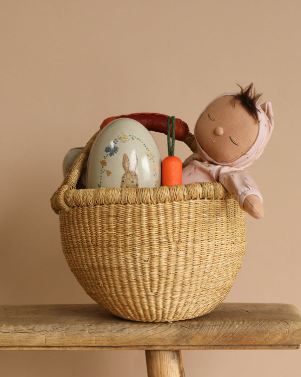 An Easter Basket Set sits on a wooden surface, featuring an Olli Ella x Odin Parker Dozy Dinkum doll with a pink headscarf, an Erzi Carrot Pretend Food toy, and an oval box with a rabbit illustration. The backdrop is soft and neutral.