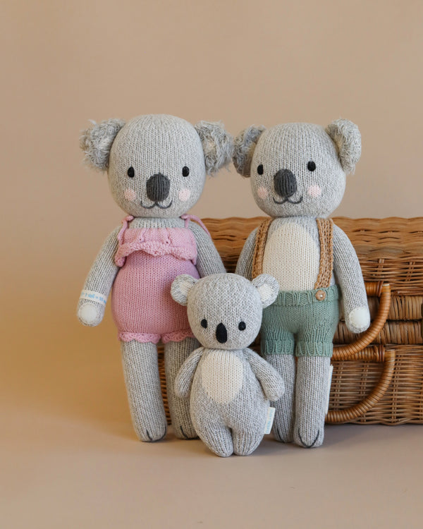 Three Cuddle + Kind Koala Stuffed Animals posed in front of a wicker basket: two large koalas, one in pink overalls, the other in green, stand behind a smaller koala in white.