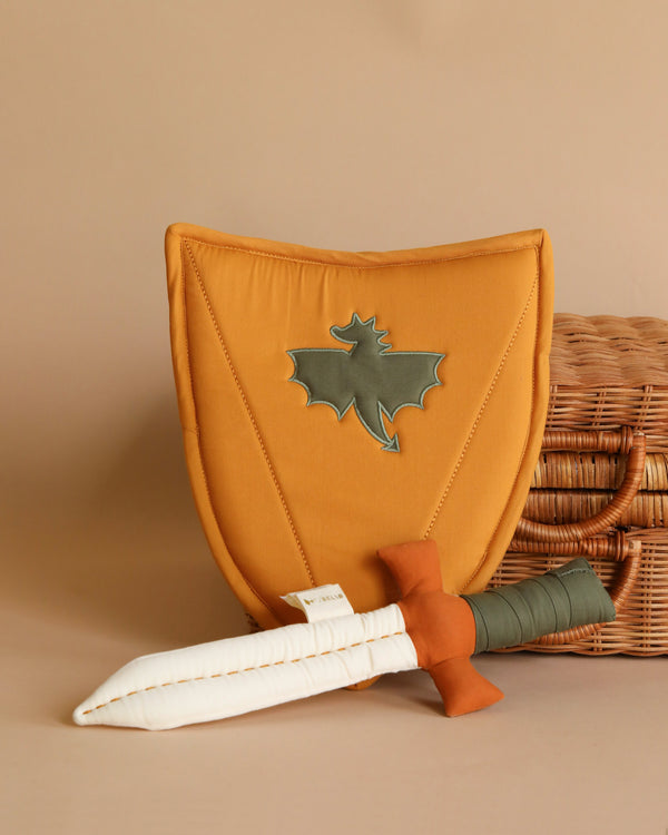 A shield and sword made out of fabric leaning against a rattan suitcase. The shield has a shape of a dragon sewn in the center.
