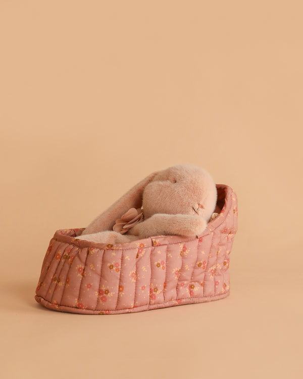The Maileg Sleeping Bunny in Carry Cot, featuring floppy ears, rests in a large pink quilted floral carrier against a plain peach background.