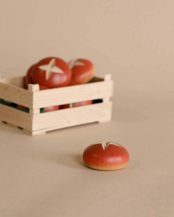A wooden crate containing Erzi Pretzel Roll pretend food, each marked with a white star, sits in the background. One tomato lies in front with a similar star marking, against a beige backdrop featuring miniature food.