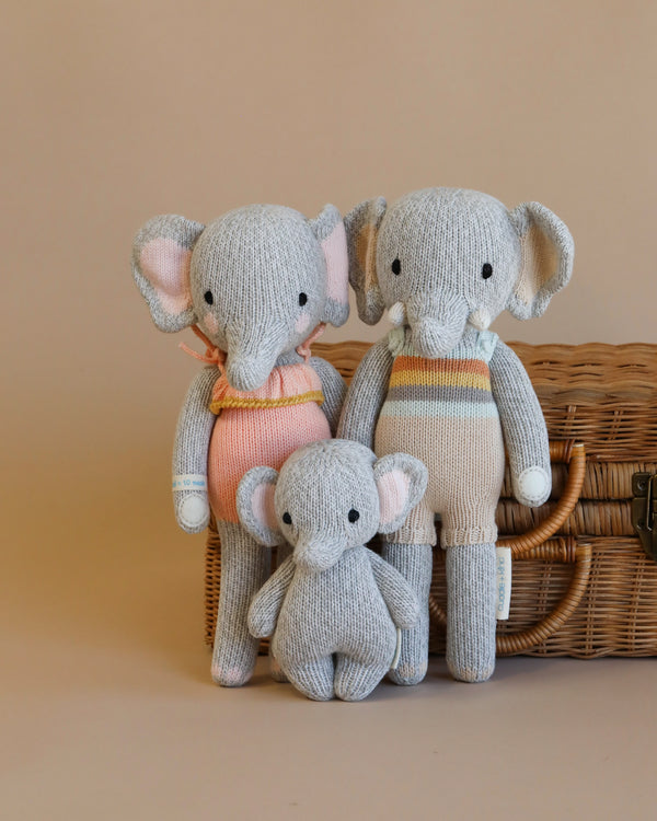 Three Cuddle + Kind Elephant Stuffed Animals - two larger and one smaller - sit in front of a wicker basket against a soft beige background. The elephants feature shades of gray with colorful clothing details.