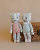 Two Cuddle + Kind Cat Stuffed Animals stand hand-in-hand in front of a wicker basket. The doll on the left wears a pink floral dress and the one on the right sports blue and white striped over