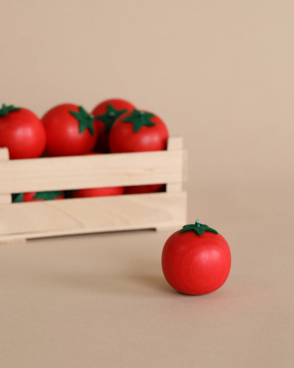 A wooden crate filled with bright red Erzi Tomato Pretend Food tomatoes stands on a soft beige background, with one tomato placed in front of the crate.
