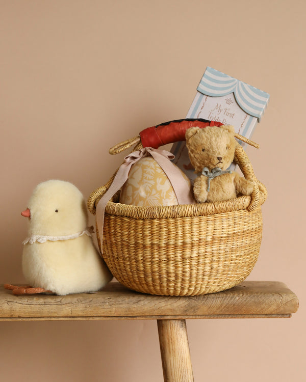 An Easter Basket, woven by hand, holds a Maileg Teddy bear, book, and fabric-wrapped item on the bench. Nearby is a plush yellow chick toy on a soft beige backdrop.