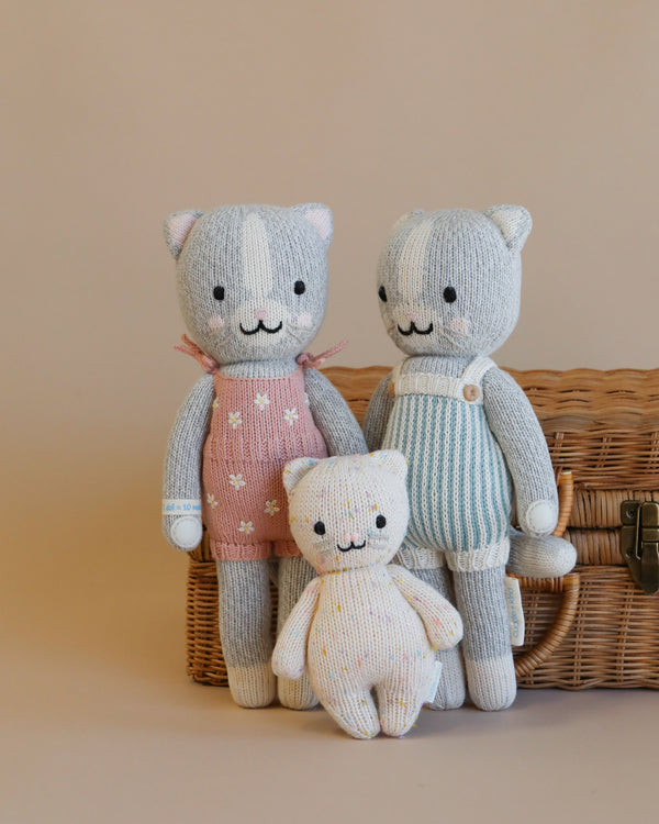 Two Cuddle + Kind Cat Stuffed Animals, one in pink and the other in blue overalls, standing next to a wicker basket with a smaller cat doll in front, against a neutral background.