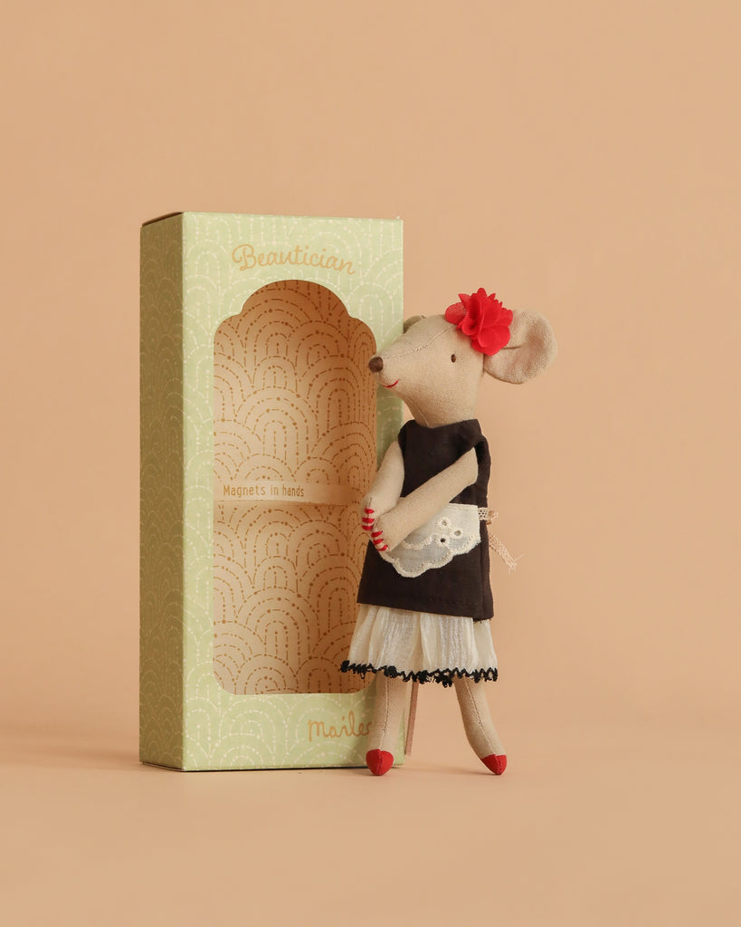 The Maileg Beautician Mouse, Mum wears a black dress, white apron, red shoes, and a red flower in its ear, standing beside a decorative green "Beautician" box on a soft beige background.