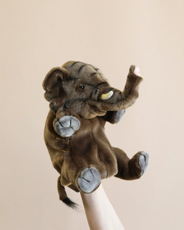 A plush toy Elephant Puppet, crafted from quality man-made materials, held up against a light beige background, featuring a raised trunk and large ears, held by a human hand visible from the wrist down.