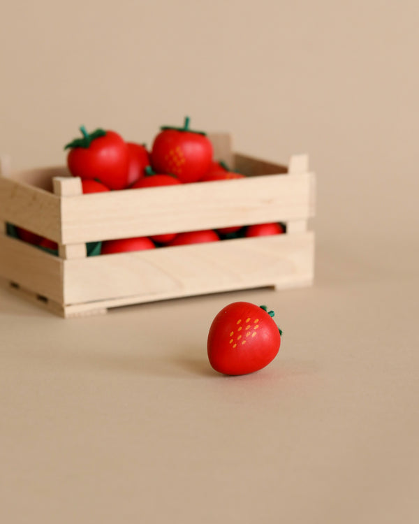 A single ripe tomato in the foreground with a wooden crate full of Erzi Strawberry Pretend Food miniature strawberries in the background, set against a neutral beige background.