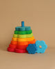 The Grimm's Flower Stacking Tower is a colorful wooden stacking toy, made from non-toxic materials, featuring various sized rings in a gradient of rainbow colors. Arranged from largest to smallest on a blue central peg, it promotes fine motor skills. Two blue flower-shaped pieces accompany the stacked rings on a beige background.