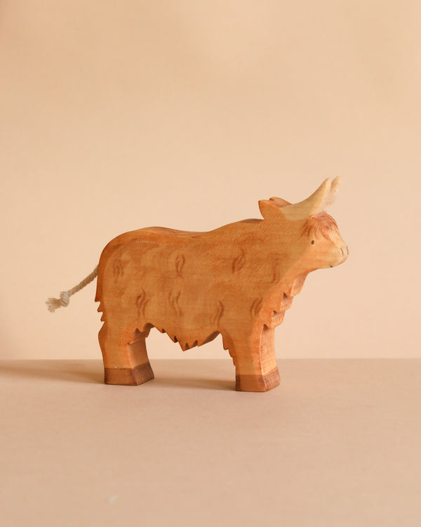 The Handmade Wooden Highland Cow, crafted from sustainable hardwood, stands on a beige surface. It features carved details, a raised head, short horns, and a rope tail against a coordinating beige background.